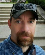 headshot photo of Dr. Giampaolo Digragorio with short dark brown hair and reddish brown mustache beard wearing a black baseball cap with black rim sunglasses on top and a blue checkered button up shirt